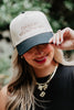 Smiling woman in a hat reading "i'm nicer when i'm tan." She wears red lipstick, a black top, layered necklaces, and is outdoors against a blurred green background.