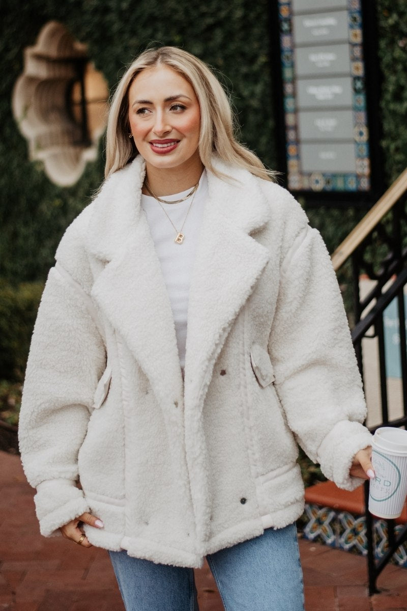 A woman with long blonde hair smiles while wearing a cozy white fleece jacket and blue jeans