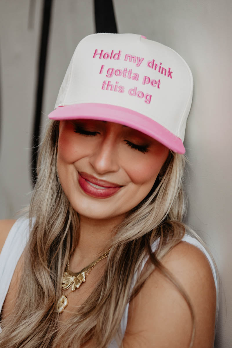 A woman wearing a pink and white two toned trucker hat that reads hold my drink I gotta pet this dog