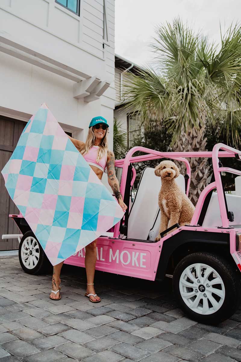 A woman and a dog in a pink jeep holding a blue and pink checkered beach towel